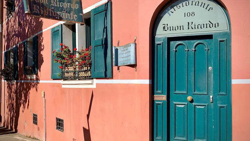 A pink building with teal windows and a door labeled “Ristorante Buon Ricordo 108.” A window box with red flowers sits beneath a sign reading “Buon Ricordo Ristorante.”.