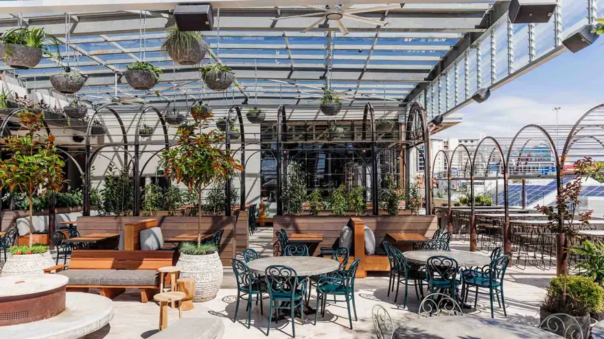 An outdoor café with many empty tables and chairs under a pergola, surrounded by greenery. 