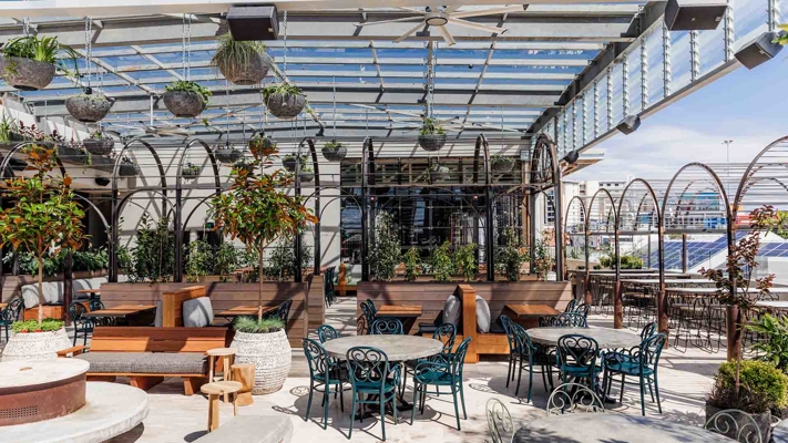 An outdoor café with many empty tables and chairs under a pergola, surrounded by greenery. 