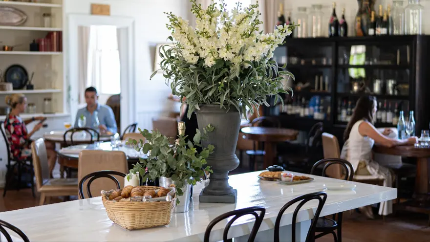 A bright, cozy café with a large floral arrangement and a basket of pastries on a white table. People are seated at tables in the background, enjoying drinks and conversation. Shelves and cabinets line the walls.