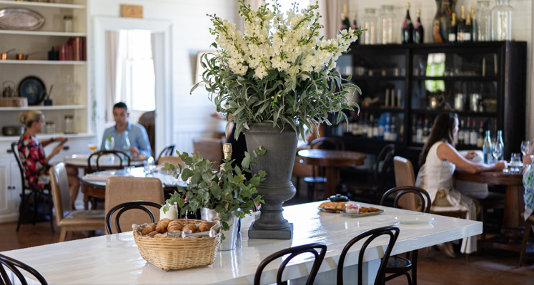 A bright, cozy café with a large floral arrangement and a basket of pastries on a white table. People are seated at tables in the background, enjoying drinks and conversation. Shelves and cabinets line the walls.